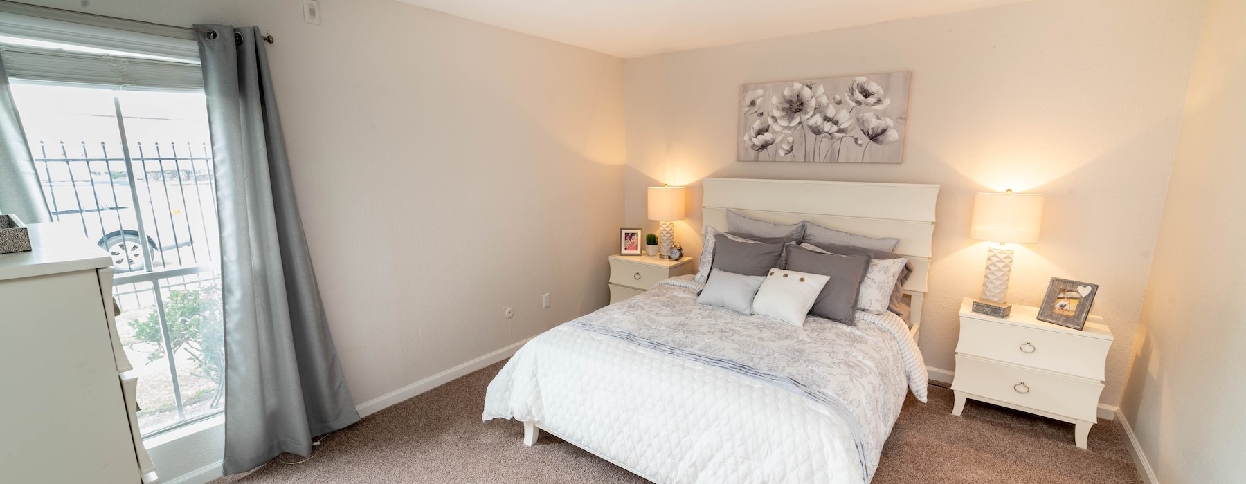 A bedroom with a large bed, two side tables with lamps on them, and a large window with curtains in a one bedroom apartment in Houston, TX at Somerset apartments.